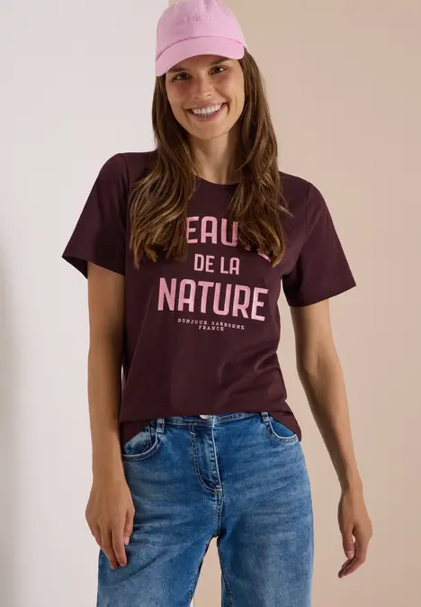 Eine Frau trägt eine rosa Cap, ein burgunderfarbenes T-Shirt mit Aufdruck und blaue Jeans, sie lächelt.