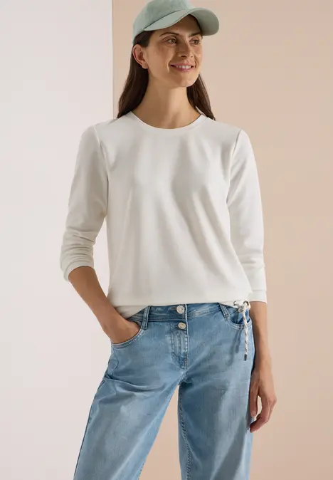 Junge Frau mit weißem Langarmshirt, blauen Jeans und Cap, lächelnd mit einer Hand in der Hosentasche.