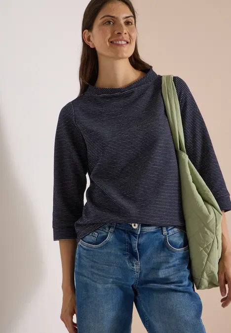 Junge Frau in Jeans und dunkelblauem Pullover trägt eine grüne Umhängetasche und lächelt.