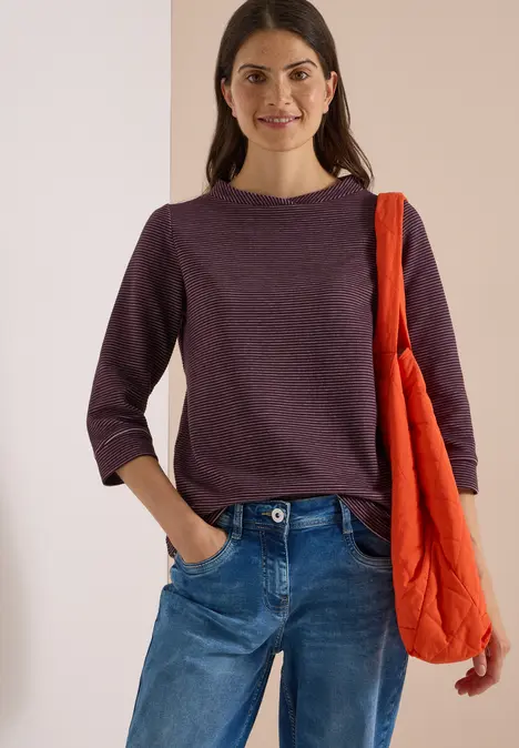 Eine lächelnde Frau in lila 3/4-Arm-Pullover und Jeans trägt eine rote Stofftasche über der Schulter.
