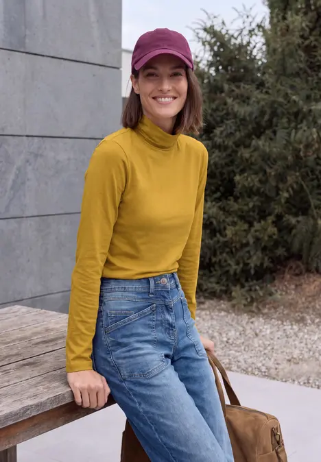 Eine Frau mit gelbem Rollkragenpullover, Jeans und pinker Mütze lehnt lächelnd an einer Holzbank.