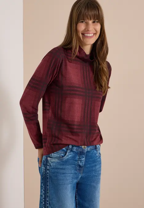 Eine junge Frau in roter karierten Bluse und blauen Jeans lächelt in die Kamera.