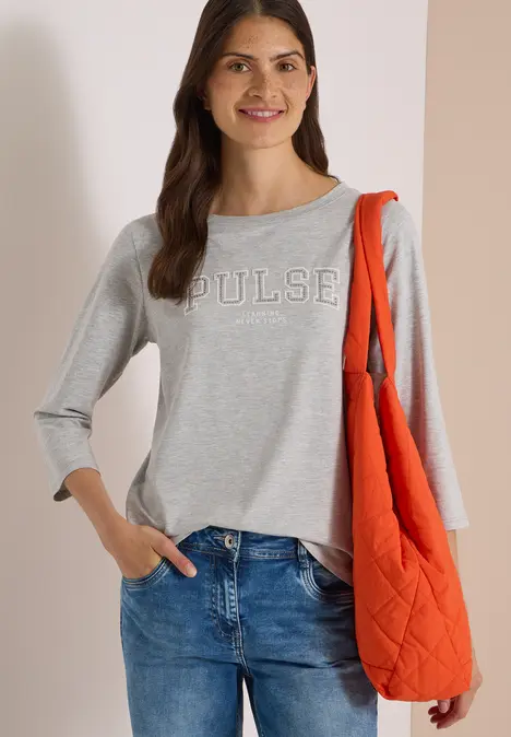 Frau trägt grauen Pullover mit 'PULSE', blaue Jeans und rote Stepp-Tasche, lächelt vor hellem Hintergrund.