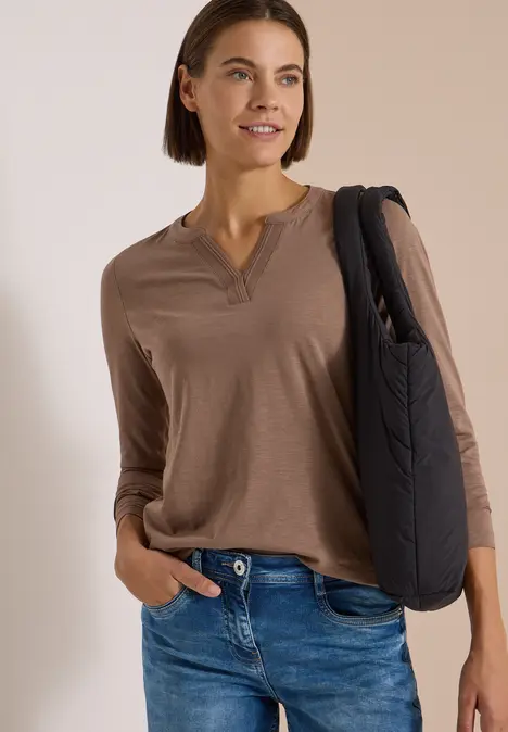 Frau mit braunem Langarmshirt, Jeans und schwarzer Schultertasche, lächelnd vor hellem Hintergrund.
