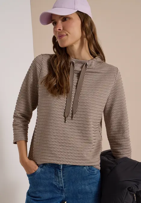 Junge Frau trägt eine strukturierte Kapuzenpullover-Bluse mit 3/4-Ärmeln, Jeans und Cap, sie hält eine Jacke.