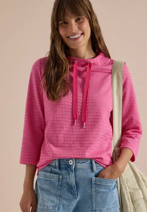 Frau trägt rosa Kapuzenpullover und Jeans, lächelnd mit Tasche über der Schulter.