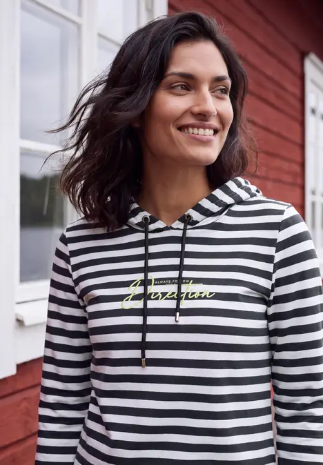 Frau im schwarz-weißen Hoodie mit gelbem Aufdruck lächelt vor roter Fassade.