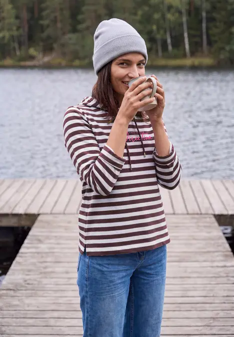 Junge Frau in gestreiftem Pullover steht auf einem Steg am See und trinkt lächelnd aus einer Tasse.