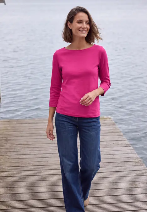Frau in pinkem Shirt und Jeans spaziert auf einem Holzsteg am Wasser, lächelnd.