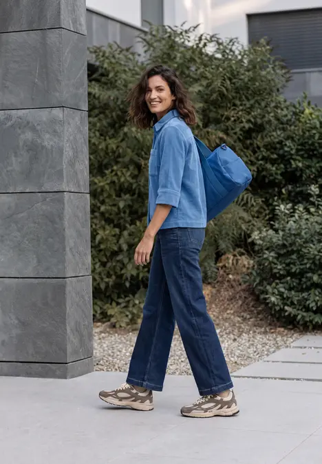 Eine junge Frau in Jeans und Sneakers mit blauem Rucksack geht entspannt auf dem Bürgersteig.