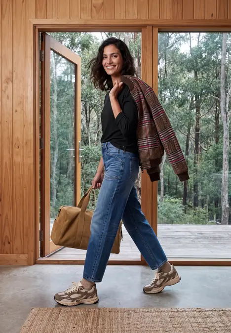 Frau in Jeans, Sneakern und Jacke über der Schulter hält Tasche und steht in einem Holzhaus vor Wald.