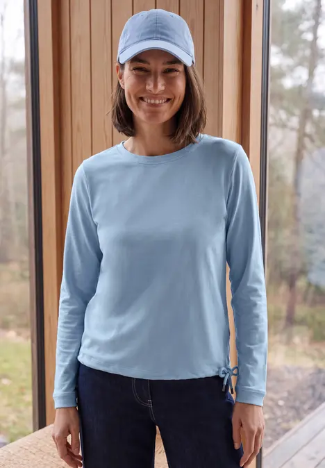 Eine Frau in Jeans, langärmeligem hellblauen Shirt und Cap lächelt vor einem Fenster mit Holzrahmen.