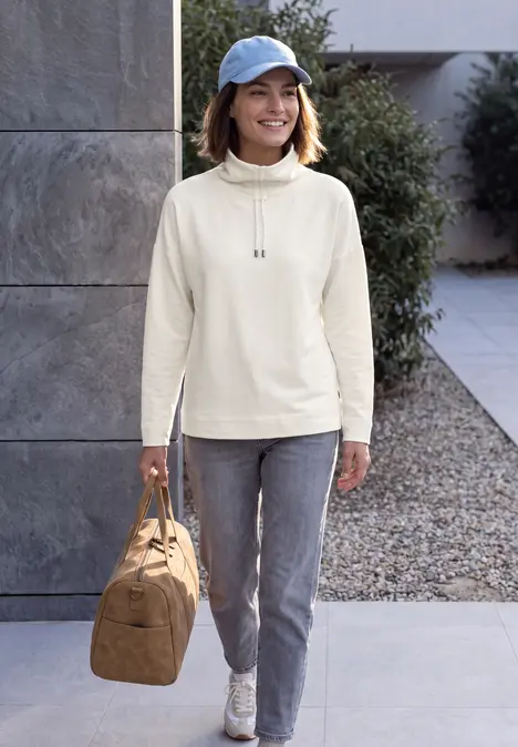 Eine Frau in heller Jacke, Jeans und Cap trägt eine Tasche und läuft vor modernem Gebäude entlang.