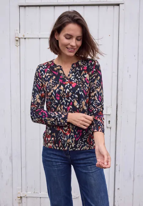 Frau in Blumenshirt und Jeans vor weißer Holztür, lächelnd und Ärmel zurechtrückend.
