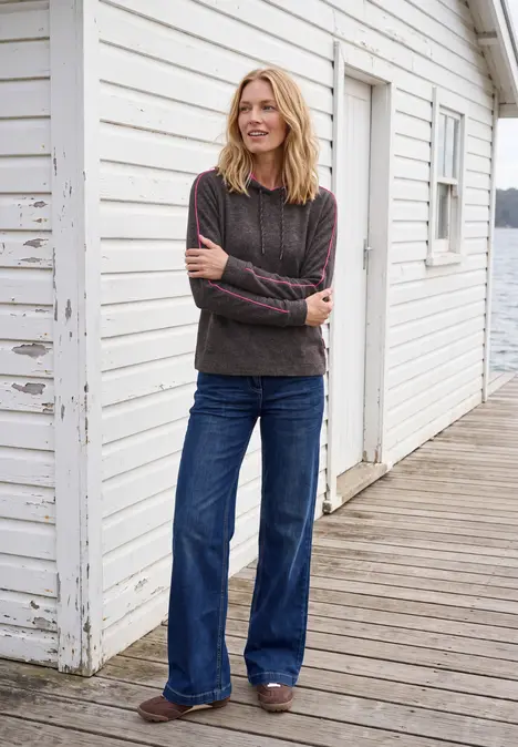 Eine lächelnde Frau im dunklen Pullover und weiten Jeans steht vor einer weißen Holzwand auf einer Veranda.