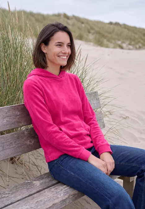 Eine Frau in pinkem Hoodie und Jeans sitzt lächelnd auf einer Holzbank am Strand vor Dünen.