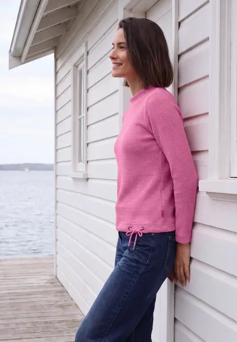 Frau in rosa Pullover lehnt an weißem Strandhaus, Blick auf ruhige See im Hintergrund.