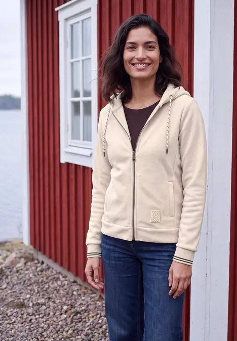 Eine junge Frau in heller Jacke und Jeans steht lächelnd vor einem roten Holzhaus am Wasser.