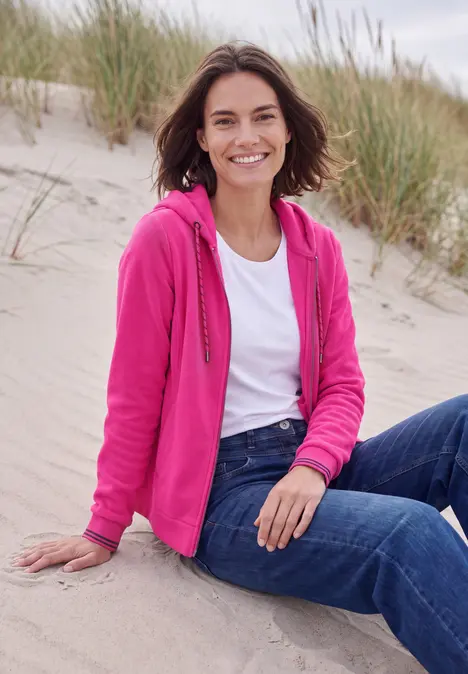 Frau sitzt lächelnd im Sand vor Dünen, trägt pinke Jacke, weißes T-Shirt und Jeans.