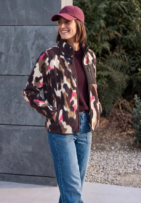 Frau mit Camouflage-Jacke, Jeans und Cap lächelt draußen vor einer Steinwand und Büschen.
