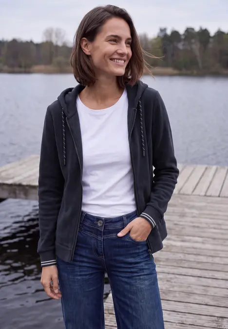 Frau im Hoodie, weißem T-Shirt und Jeans steht lächelnd auf einem Steg am See.