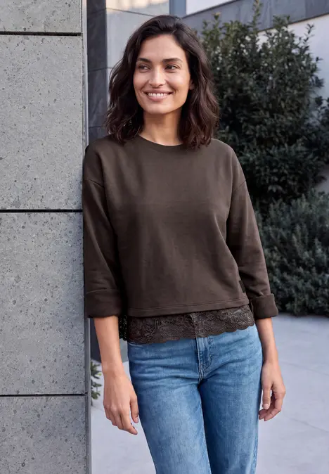 Eine lächelnde Frau mit braunem Pullover und blauen Jeans lehnt an einer Mauer im Freien.