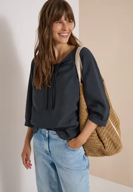 Frau in dunkler Kapuzenbluse und hellen Jeans mit Korbtasche auf dem Arm, lächelt vor hellem Hintergrund.