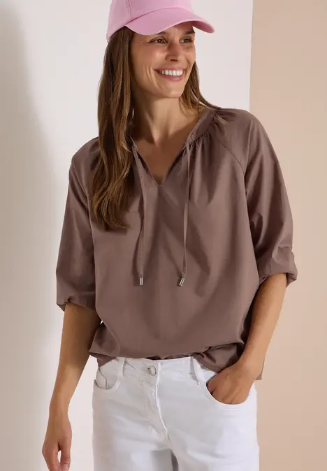 Eine lächelnde Frau mit rosa Cap, lockerer brauner Bluse und weißen Hosen, Hand in Tasche.