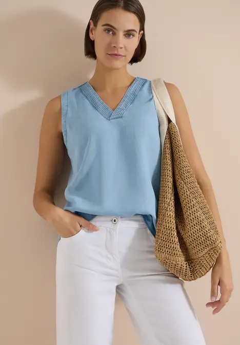 Frau in hellblauem ärmellosen V-Ausschnitt-Top, weißen Hosen und großer Strohtasche vor neutralem Hintergrund.