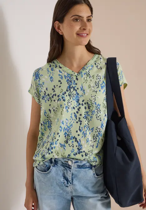 Frau in geblümter grüner Bluse und Jeans trägt lächelnd eine blaue Tasche über der Schulter.