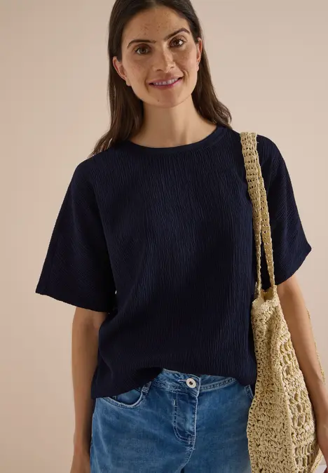 Eine lächelnde Frau im dunkelblauen Shirt, Jeans und einer Netztasche vor hellem Hintergrund