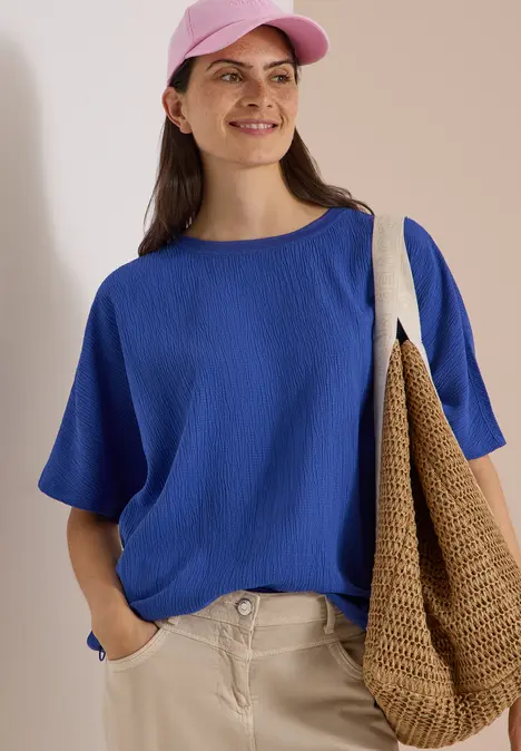 Frau in blauer Bluse, rosa Hut und beiger Hose, lächelt und trägt eine geflochtene Tasche.
