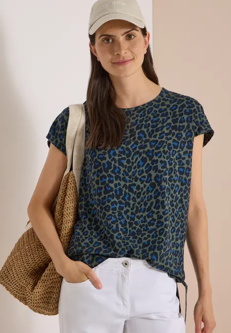 Frau in blau-grünem Leo-Shirt, weißer Hose und Strohtasche, mit Cap, lässig lächelnd