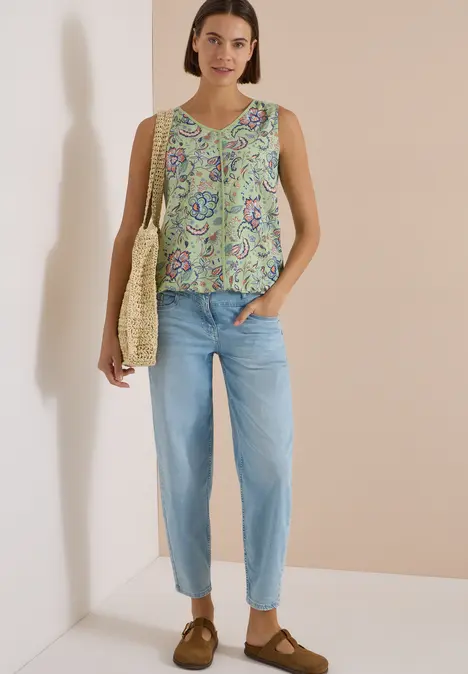 Eine Frau trägt ein ärmelloses, geblümtes Top, hellblaue Jeans, braune Schuhe und eine Korbtasche.