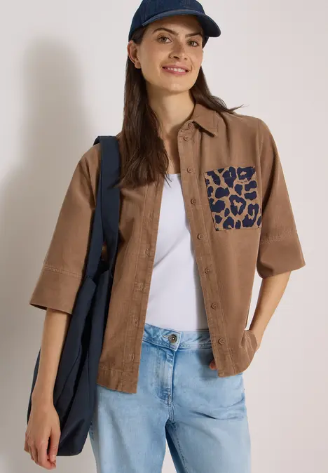 Junge Frau in braunem Kurzarmhemd mit Leoparden-Tasche, weißem Top, Jeans, navy Cap und Tasche.