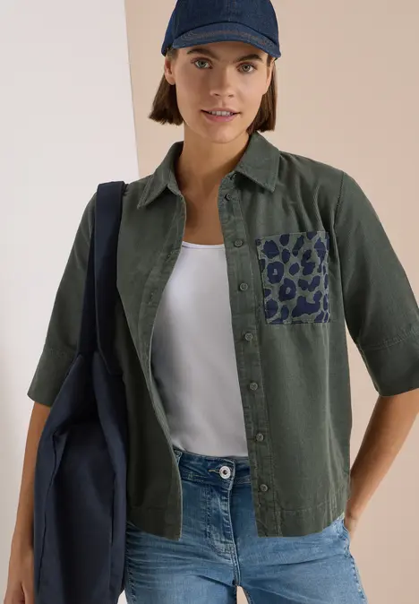 Frau trägt grüne Bluse mit Leopardenmuster-Tasche, weißem Top, Jeans, dunkelblauer Cap und Tasche.