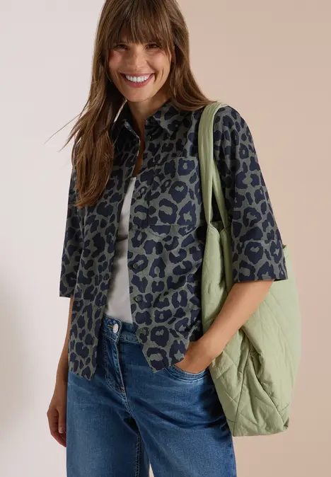 Eine lächelnde Frau trägt eine gemusterte Jacke, weißes Shirt, blaue Jeans und grüne Tasche.