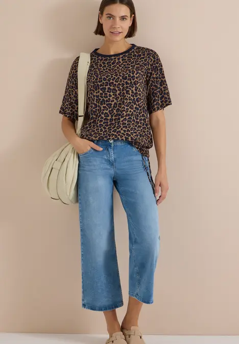 Eine Frau trägt eine Leo-Bluse, weite Jeans-Culotte und helle Tasche vor beigem Hintergrund.