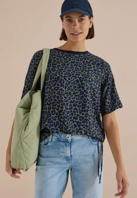Frau in weitem, blau-grün gemustertem T-Shirt, Jeans, dunkelblauer Mütze und großer, grüner Umhängetasche.