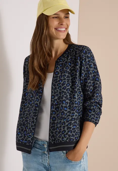 Lächelnde Frau mit blauer Leo-Bomberjacke, weißem Top, Jeans und gelber Kappe.
