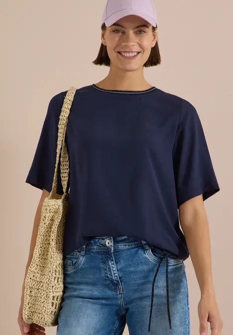 Lächelnde Frau in dunkelblauem T-Shirt, Jeans und Baseballcap trägt eine geflochtene Umhängetasche.
