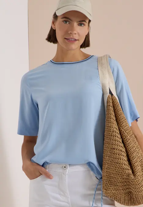 Frau in hellblauem T-Shirt, weißer Hose und Cap, mit Strohtasche vor hellem Hintergrund.