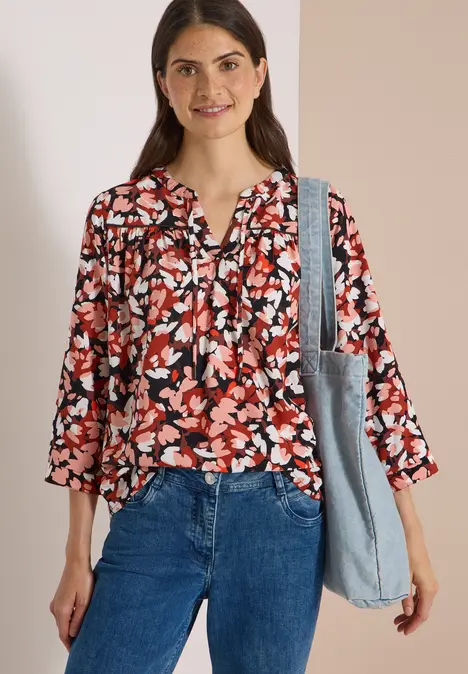 Frau in floraler Bluse und Jeans, lächelt und trägt eine Schultertasche vor hellem Hintergrund.