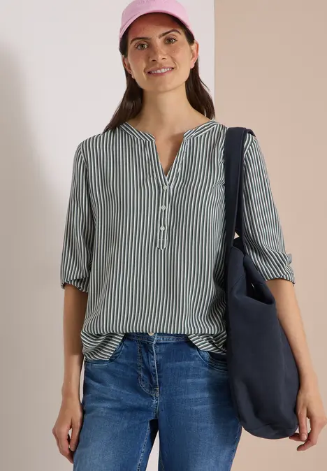Frau in grün-weiß gestreifter Bluse, Jeans, rosa Cap und Umhängetasche, stehend und lächelnd.
