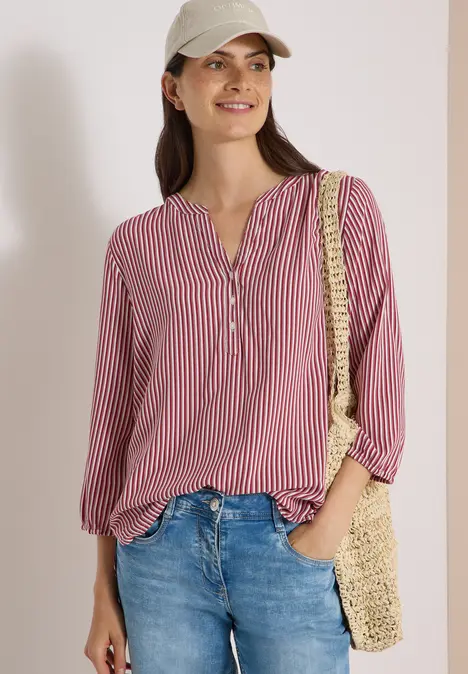 Frau mit roter, gestreifter Bluse, Jeans, Strohtasche und Cap, lächelnd vor weißem Hintergrund.