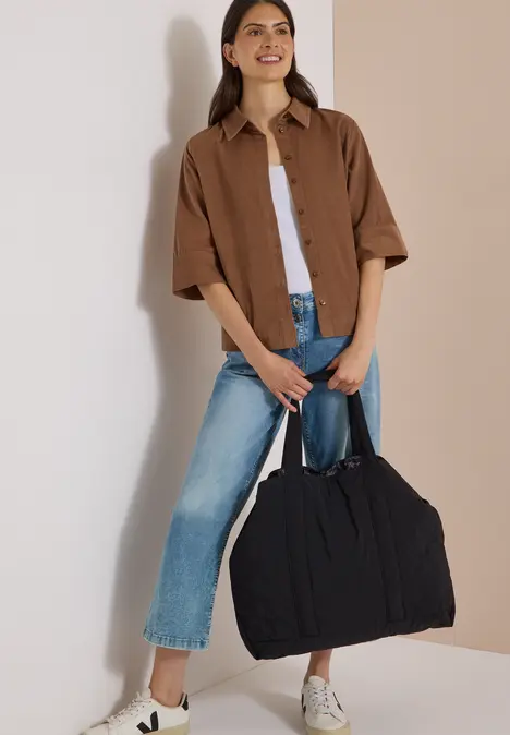 Frau im Outfit mit Jeans, weißem Top, brauner Hemdbluse und weißen Sneakern, die eine große schwarze Tasche trägt.