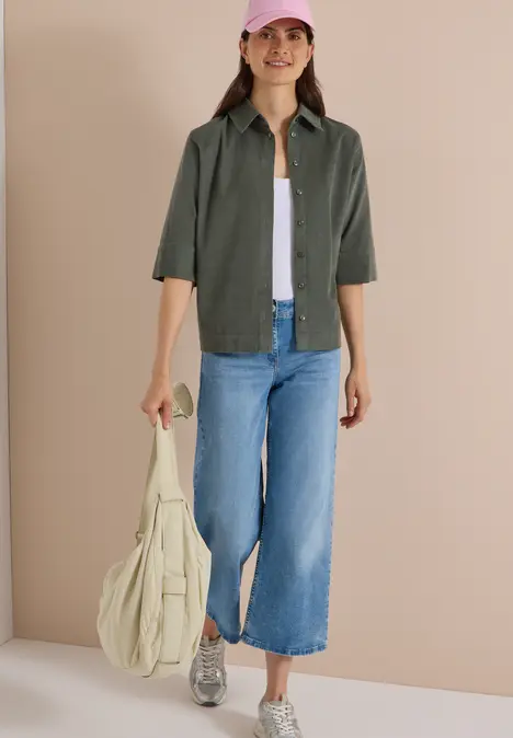 Frau in weitem Jeans-Culotte, olivgrünem Hemd, weißem Top, Sneakern und pinker Kappe trägt Tasche.