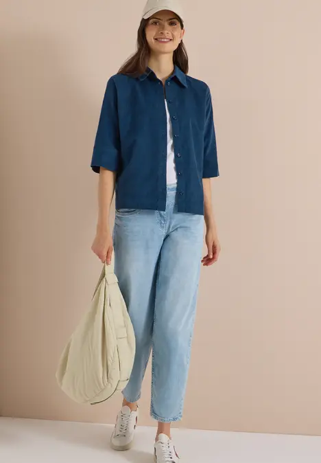 Eine lächelnde Frau trägt Jeansjacke, Jeans, weiße Sneakers, Baseballcap und hält eine große beige Tasche.
