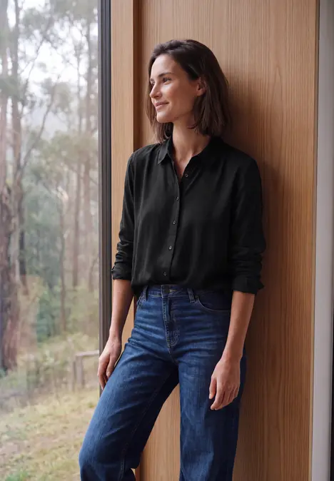 Eine lächelnde Frau in schwarzem Hemd und Jeans lehnt an einer Holzwand mit Blick ins Grüne.