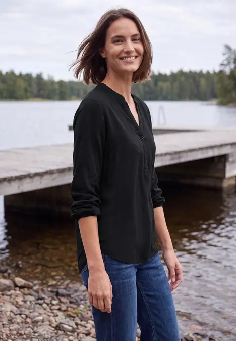 Eine Frau in schwarzer Bluse und Jeans steht lächelnd an einem Steg an einem ruhigen See.
